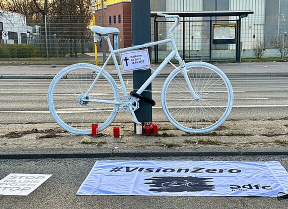 Geisterrad Neukölln 2025 ein weißes Geisterrad mit Informationsschild "Radfahrer 63 Jahre" und Vision Zero Fahne im Vordergrund