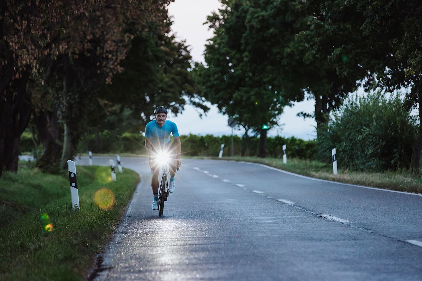 Sigma Landstraße Ein Rennradfahrer fährt in der Dämmerung auf einer Landstraße auf die Kamera zu, am Lenker ein Scheinwerfer. Im Hintergund grüne Landschaft.